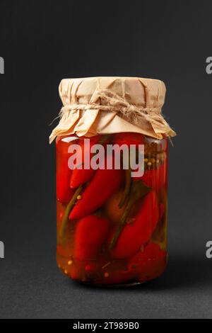 Eingelegte rote Chilischoten in einem geschlossenen Glasgefäß, isoliert auf schwarzem Hintergrund. Gemüse aus der Dose. Scharfer Snack. Stockfoto
