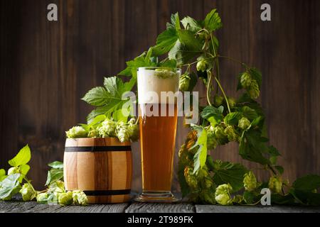 Ein Glasbecher Bier, ein Hopfenzweig und ein Holzfass mit Hopfen auf dunklem hölzernem Hintergrund. Brautraditionen. Oktoberfest. Bierfest. Stockfoto