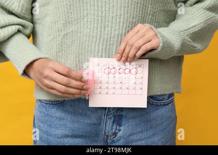 Junge Frau mit Menstruationstasse und Kalender mit markierten Menstruationstagen auf gelbem Hintergrund Stockfoto