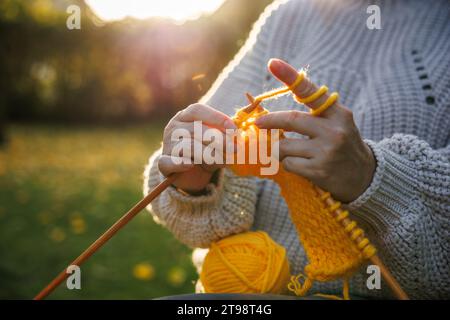 Hände stricken mit Wolle. Frau im Wollpullover sitzt draußen und strickt warme Kleidung aus gelbem Garn Stockfoto