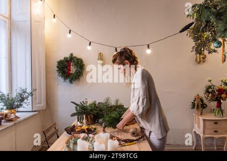 Die junge Frau macht zu Hause einen handgefertigten Weihnachtskranz aus natürlichen Materialien. Authentisches Apartment mit skandinavischem Interieur. DIY-Anweisung von flori Stockfoto