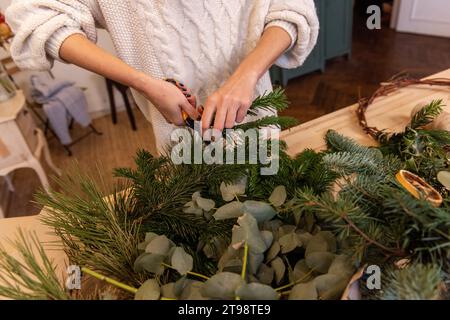 Draufsicht aus weiblichen Händen, die natürliche Zweige von Fichte, Kiefer, Eukalyptus mit Garn an die botanische Basis binden. Das Mädchen macht einen Adventsgott Stockfoto