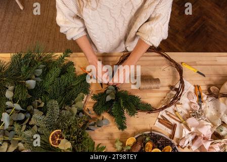 Draufsicht Komposition weiblicher Hände, die Weihnachtskranz aus natürlichen Zweigen von Fichte, Kiefer, Eukalyptus herstellen. Das Mädchen wickelt grüne Zweige auf die Kraft-Basis Stockfoto