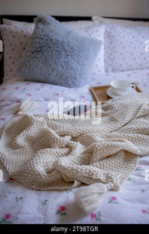 Ein gemütliches Innenleben im Schlafzimmer mit einem Buch, einer Tasse und einer weißen Strickdecke auf einem Bett Stockfoto