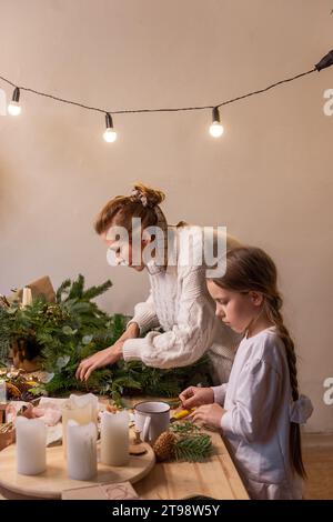 Junge Mutter und kleine Tochter machen zusammen Weihnachtskranz aus Fichte, Eukalyptuszweigen. Alleinerziehende Mutter zeigt Mädchen Tutorial, wie man Neues macht Stockfoto