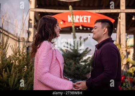 Verliebtes Paar wählt Weihnachtsbaum auf dem Markt im Freien. Ein junger Mann und eine schöne Frau sehen sich an, halten eine Wanne mit einem lebendigen Weihnachtsbaum in han Stockfoto