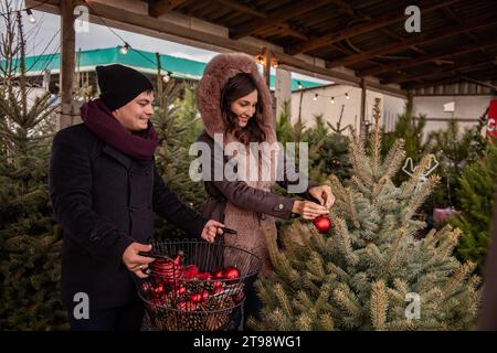 Lustiges Paar schmückt Weihnachtsbäume auf dem Markt mit roten Kugeln. Junge Männer und Frauen hängen Spielzeug an Tannenbäumen im Garten des Hinterhofs auf, um gemütlich und festlich zu gestalten Stockfoto