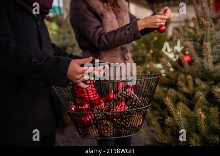 Nahaufnahme der Hände des Mannes, die einen Eisenkorb mit roten Kugeln halten. Defokussierter Hintergrund die Mädchenhände schmücken den Weihnachtsbaum. Weihnachtliche Atmosphäre Stockfoto