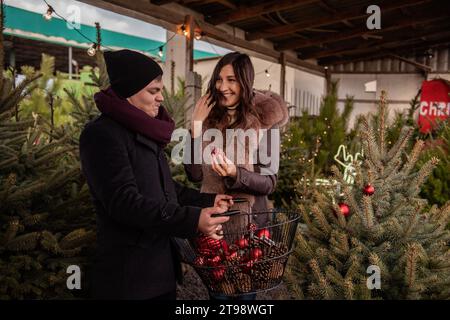 Das lustige Paar macht sich rumlustig und schmückt den Weihnachtsbaum auf dem Markt mit roten Kugeln. Junge Mann und Frau hängen Spielzeug an Tanne im Garten, Crea Stockfoto