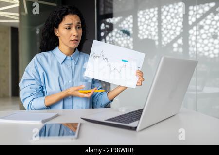 Verärgerte und verwirrte Frau bei Online-Videobesprechung zeigt Kollegen Partner, Diagrammberichte mit negativen Ergebnissen, Geschäftsfrau im Büro hinter Papierarbeit. Stockfoto