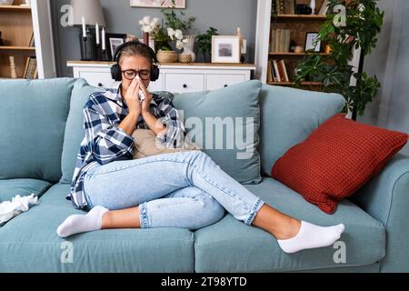 Junge traurige Frau, die allein zu Hause auf dem Sofa sitzt und traurige Musik auf kabellosen Kopfhörern hört, weint und wischt sich mit einem Papiertaschentuch die Nase ab. Erinnerungen an Stockfoto