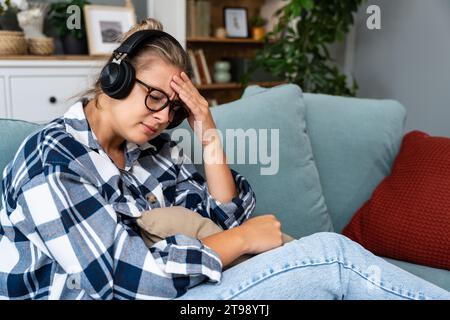 Junge traurige Frau, die allein zu Hause auf dem Sofa sitzt und traurige Musik auf kabellosen Kopfhörern hört, weint und wischt sich mit einem Papiertaschentuch die Nase ab. Erinnerungen an Stockfoto