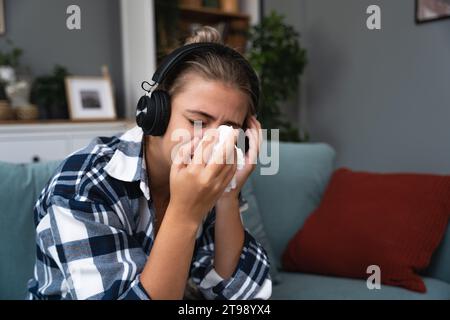 Junge traurige Frau, die allein zu Hause auf dem Sofa sitzt und traurige Musik auf kabellosen Kopfhörern hört, weint und wischt sich mit einem Papiertaschentuch die Nase ab. Erinnerungen an Stockfoto