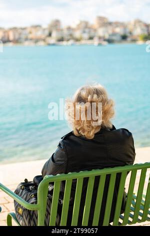 Alter - allein. Alte Frau mit Tasche, die von hinten gesehen wird und auf das Meer blickt. Großmutter Einsamkeit. Rentner sitzt auf einer Bank, Stockfoto