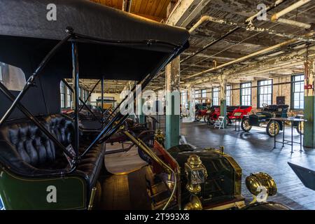 Der offene Vordersitz einer historischen Kutsche mit schwarzem Klappdach und Messinglaternen ist im Vordergrund sichtbar, dahinter sind Fahrzeuge in der Fabrikhalle des Ford Piquette Plant in Detroit, Michigan, USA, aufgereiht. Erbaut 1904 gilt das Ford Piquette Plant als das älteste erhaltene Gebäude, das ursprünglich für die Autoproduktion errichtet wurde und das der Öffentlichkeit zugänglich bleibt. Stockfoto