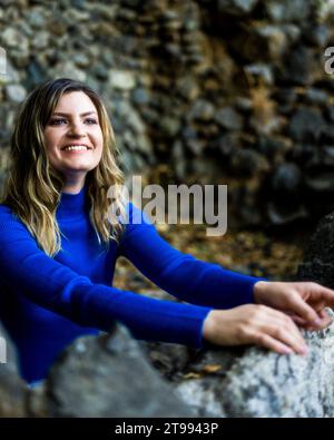 Junge Frau mit blauem Langarm-Crop-Top und weißer Denim-Jeans in einem von Old Rock umgebenen Brunnen Stockfoto