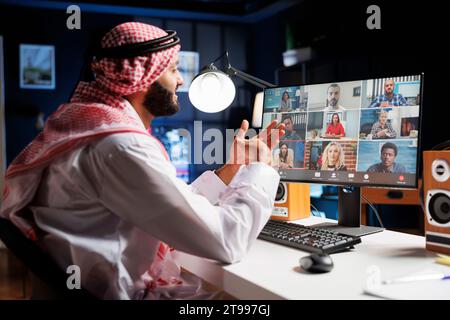 Bild eines Mannes aus dem Nahen Osten, der traditionelle Kleidung trägt und mit einer Gruppe von Menschen auf seinem Computerbildschirm spricht. Ein islamischer Kerl, der sich mit seinem Team bei einer Telefonkonferenz unterhält. Stockfoto