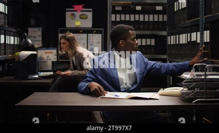 Ein männlicher Spezialist für Strafrecht untersucht Beweise und verwendet Zeugenaussagen und geheime Berichte, um den Fall zusammenzustellen. Der afroamerikanische Polizist verwendet Papierdatenbanken zur Identifizierung. Stockfoto