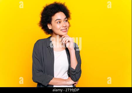 Wunderschöne lockige afroamerikanische stilvolle positive Frau, schaut zur Seite auf leeren Raum für Werbung, denkt, steht auf isoliertem gelben Hintergrund, lächelt Stockfoto