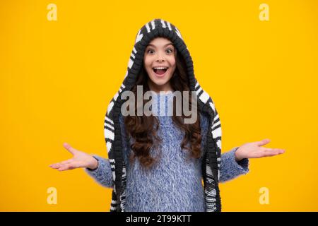 Verblüffter Teenager. Wunderschönes Winterporträt für Kinder. Teenager-Mädchen posiert mit Winterpullover und Strickmütze auf gelbem Hintergrund. Warme Mütze mit Kapuze und Stockfoto