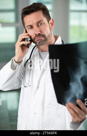 Ernsthafter Arzt, der sich Röntgen anschaut und telefoniert Stockfoto