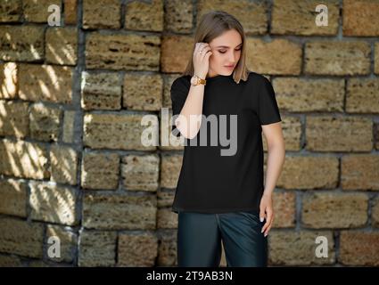 Mockup eines schwarzen T-Shirts auf einem posierenden Mädchen, auf dem Hintergrund einer Backsteinmauer mit Schatten, für Branding, Design. Hemdschablone für eine Frau, Presenta Stockfoto