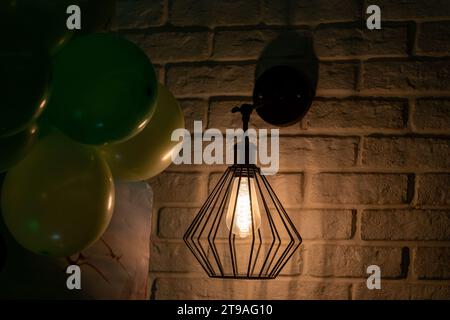 Glühbirne, an der weißen Wand. Stimmung. Atmosphäre. Hell im Dunkeln. Geburtstagsballons in der linken Ecke. Stockfoto