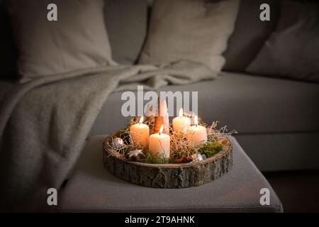 Vier weiße Adventskerzen und Weihnachtsdekoration in einem rustikalen Baumtablett auf einer Ottomane auf einer Couch, gemütliche Wintereinrichtung im Wohnzimmer, Copy Spa Stockfoto