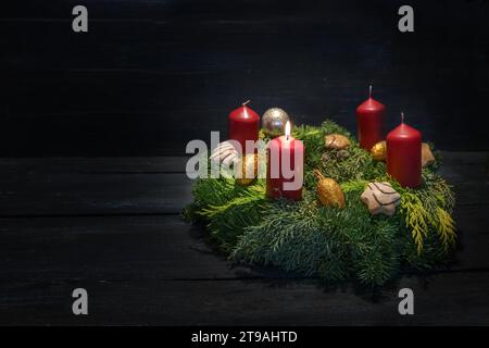 Licht im Dunkeln am ersten Advent, naturgrüner Kranz mit roten Kerzen, einer brennt, Weihnachtsdekoration und Kekse, dunkler Holzhintergrund, Co Stockfoto