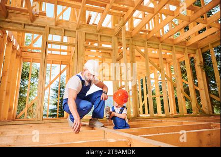 Vater mit Kleinkind-Sohn, der ein Holzrahmenhaus baut. Ein Baumann trainiert seinen Sohn, wie man einen elektrischen Schraubendreher auf der Baustelle an sonnigen Tagen bedient. Zimmerei und Familienkonzept. Stockfoto