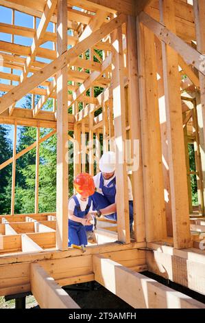 Vater mit Kleinkind Sohn baut ein Holzrahmenhaus. Der Baumeister lehrte seinen Sohn in der Kunst des Hämmerns von Nägeln auf der Baustelle und trug Helme und Overalls an sonnigen Tagen. Zimmerei und Familienkonzept Stockfoto