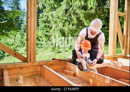 Vater mit Kleinkind-Sohn baut ein Holzrahmenhaus in der Nähe des Waldes. Männlicher Arbeiter, der seinem Sohn beibringt, wie man Bretter mit der elektrischen Säge auf der Baustelle schneidet, mit Helmen und Overalls an sonnigen Tagen. Stockfoto