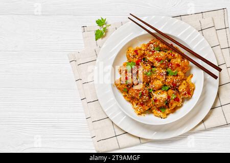 Knusprige orange Blumenkohlröschen, bestreut mit Sesamsamen, Chili-Flocken und Petersilie in weißer Schüssel auf weißem Holztisch mit Essstäbchen, horizonta Stockfoto