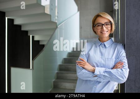 Porträt einer jungen schönen Frau am Arbeitsplatz im Büro, blonde Geschäftsfrau mit überkreuzten Armen und Brille lächelnd und in die Kamera schauend, weibliche Arbeiterin in der Nähe des Fensters. Stockfoto