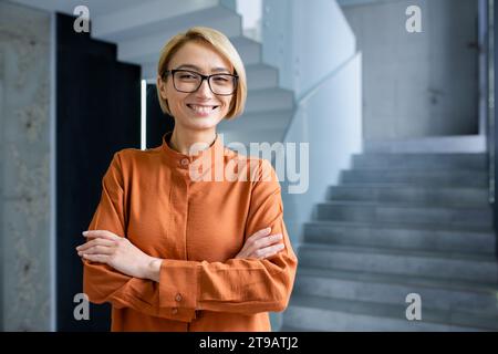 Porträt einer jungen schönen Frau am Arbeitsplatz im Büro, blonde Geschäftsfrau mit überkreuzten Armen und Brille lächelnd und in die Kamera schauend, weibliche Arbeiterin in der Nähe des Fensters. Stockfoto