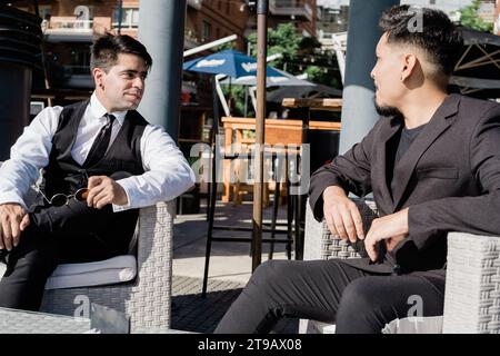 Junge lateinamerikanische Männer in eleganten Kleidern sitzen draußen in Sesseln und unterhalten sich nach dem Büro beiläufig. Stockfoto