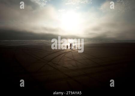 Silhouette eines männlichen Kite Buggers (Kite Buggy), der in unheilvollen Wolken und ungewöhnlichem Licht mit dunklen Spuren im Sand den Strand hinunterrollt. Stockfoto