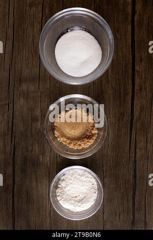 Zucker, brauner Zucker und Mehl in Glasschalen gegen Holz Stockfoto