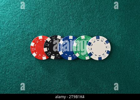 Casino-Hintergrund mit Details von fünf verschiedenen farbigen Plastikchips in einer Reihe auf grüner Filz Spielmatte. Draufsicht. Stockfoto