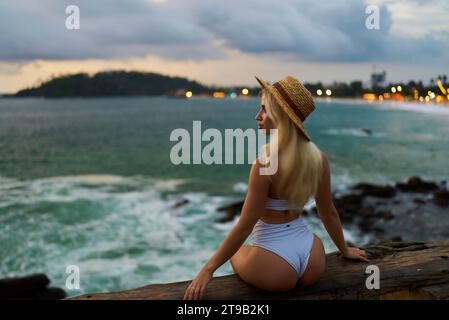 Die blonde Frau in weißem Bikini und Strohhut sitzt in der Abenddämmerung auf der Klippe am Meer, blickt über turbulente Wellen, verkörpert Alleinreisen und ruhige Besinnung Stockfoto