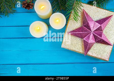 Goldene glänzende Box mit rosa Stern, brennende Kerzen mit Feuer und Kiefernkegel, Tannenzweige auf blauen Brettern. Weihnachten, Neujahr, Geburtstag, Feiertag. Kopie sp Stockfoto