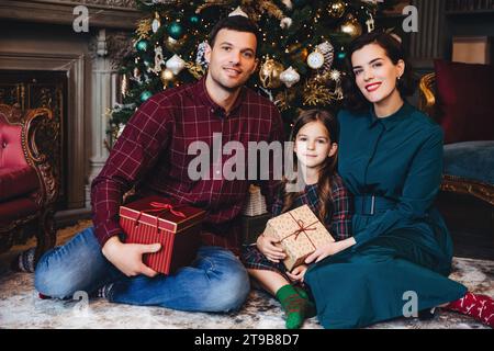 Lächelnde Familie mit einem jungen Mädchen, das Weihnachtsgeschenke hält, in einem opulenten Raum mit einem reich verzierten Baum Stockfoto