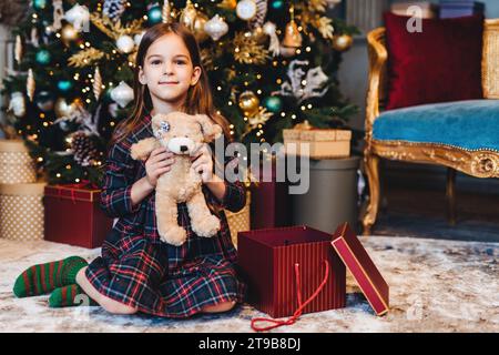 Zufriedenes Mädchen im karierten Pyjama mit einem Teddybären, einem Weihnachtsbaum und Geschenke in einem reich verzierten Zimmer Stockfoto