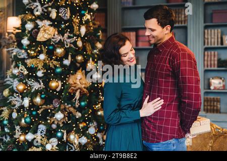 Liebevolles Paar in festlicher Kleidung, das sich vor einem wunderschön geschmückten Weihnachtsbaum umgibt und einen zarten Moment teilt Stockfoto