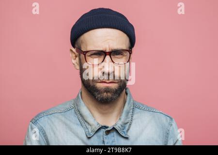 Verwirrter Mann mit Bart und Brille, der eine Mütze und ein Jeanshemd auf rosa Hintergrund trägt Stockfoto