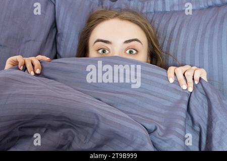 Ein Teenager mit offenen Augen bedeckt morgens ihr Gesicht mit blauer Decke. Schöne Frau, die im Bett liegt und schläft. Schlafen Sie nicht genug Stockfoto