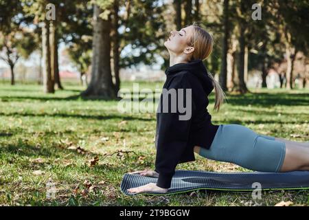 Die junge Frau mit blonden Haaren und Sportbekleidung, die im Park auf einer Yoga-Matte meditiert und sich ausdehnt Stockfoto