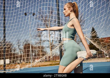 Schöne junge Sportlerin mit Pferdeschwanz, die in Aktivkleidung in der Nähe des Netzes draußen trainiert, das Bein dehnt Stockfoto