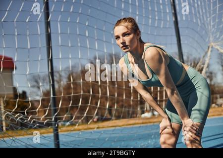 Sportliche junge Frau mit Pferdeschwanz in Aktivkleidung, die beim Training im Freien in der Nähe des Netzes ruht Stockfoto