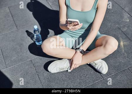 Verkürzte Passform Frau in Aktivkleidung mit Smartphone und in der Nähe von Wasserflasche sitzen, Gesundheit und Fitness Stockfoto